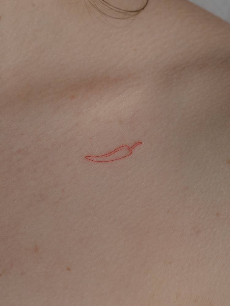 Tiny minimalist red chili pepper tattoo placed on the collarbone.