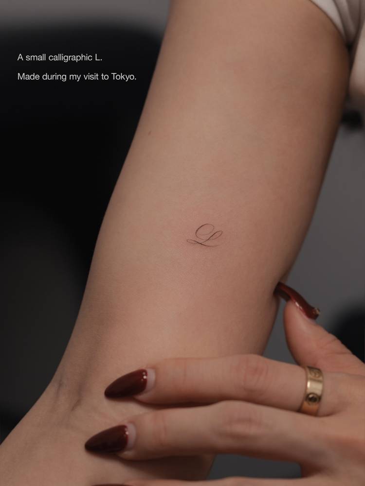 Little script font letter "L" tattoo located on the inner arm.